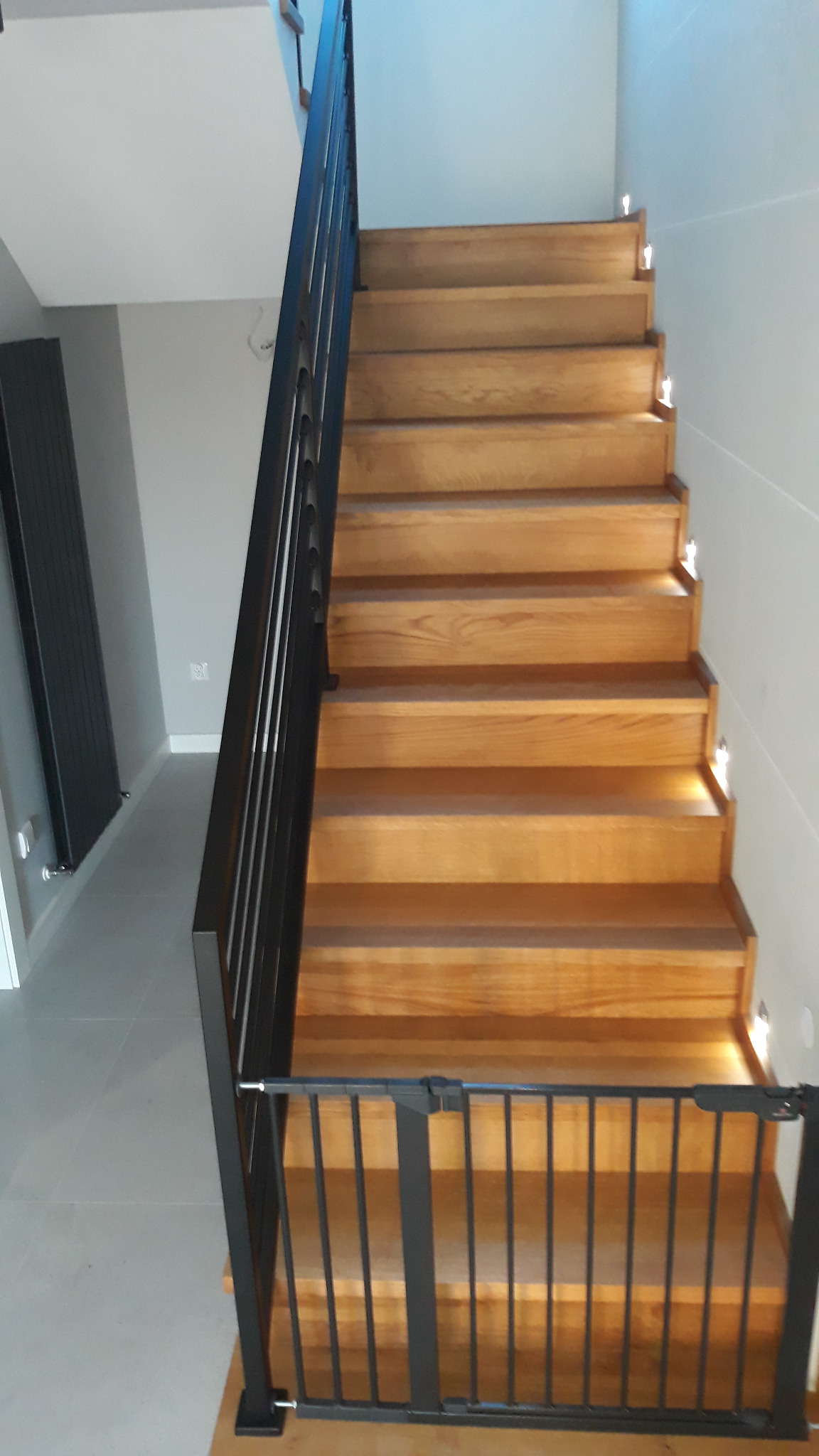 Drewniane schody z czarną balustradą i oświetleniem LED w stopniach, zabezpieczone bramką dla dzieci. Widok z dołu, nowoczesny design.