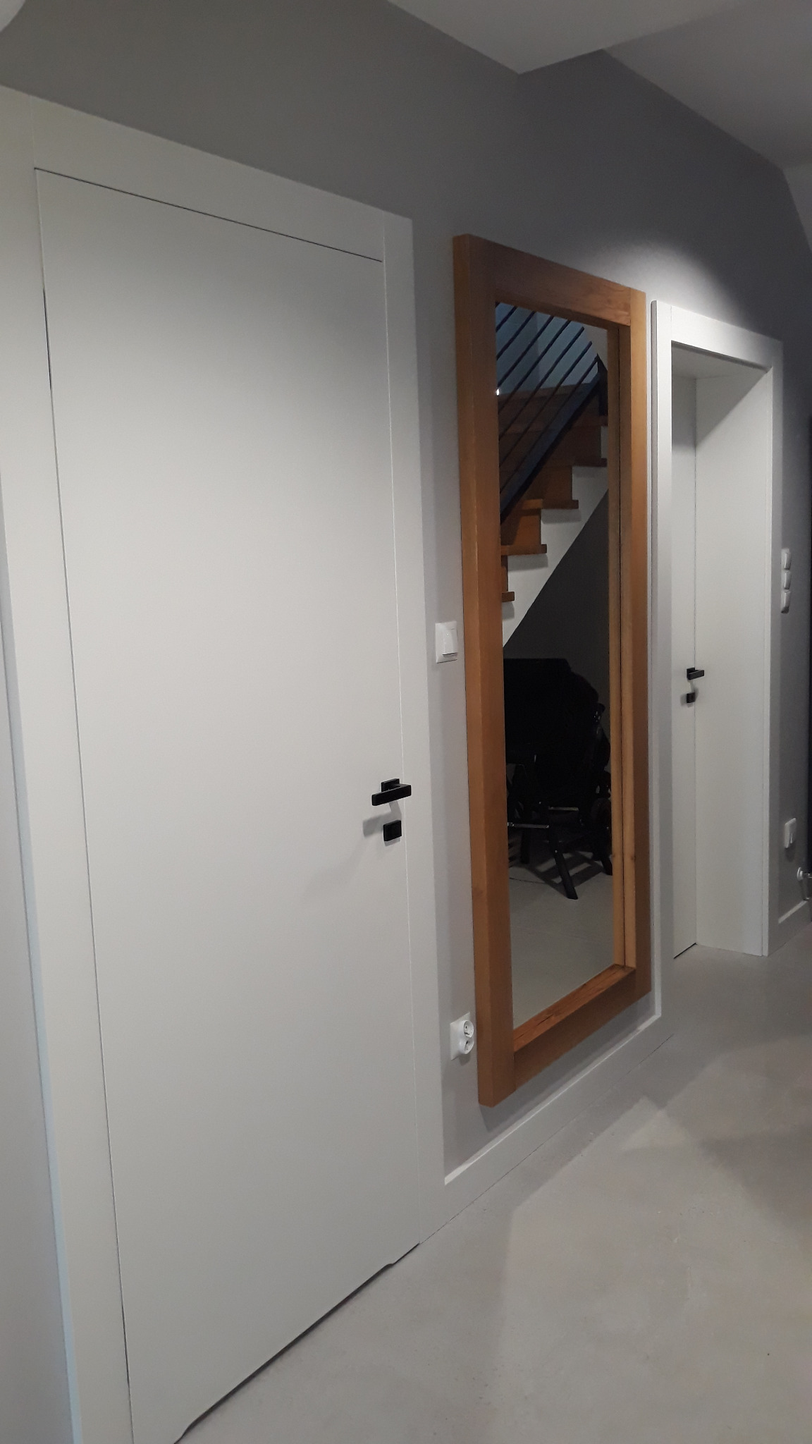 Białe, minimalistyczne skrzydło drzwiowe z czarną klamką, obok lustro w drewnianej ramie odbijające schody. W tle widoczne kolejne drzwi.