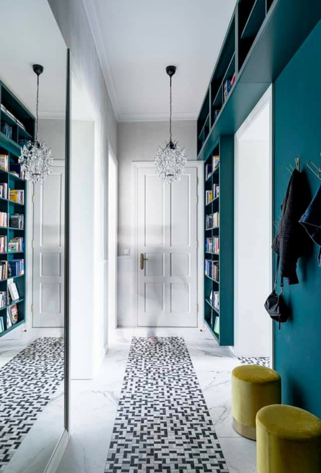 Korytarz z zabudową biblioteczną w kolorze morskim, lustrem i wzorzystą posadzką. Kryształowy żyrandol i dwa pufy dopełniają aranżację.