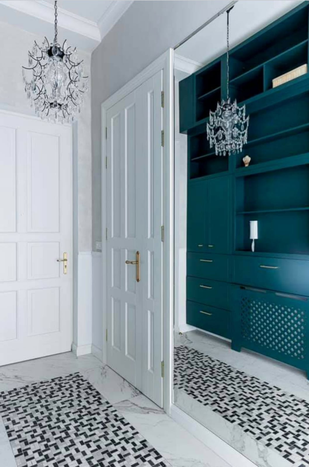 Białe drzwi wewnętrzne z złotą klamką, otwarte na szafę wnękową w odcieniach zieleni, z kryształowym żyrandolem. Marmurowa podłoga z geometrycznym wzorem.