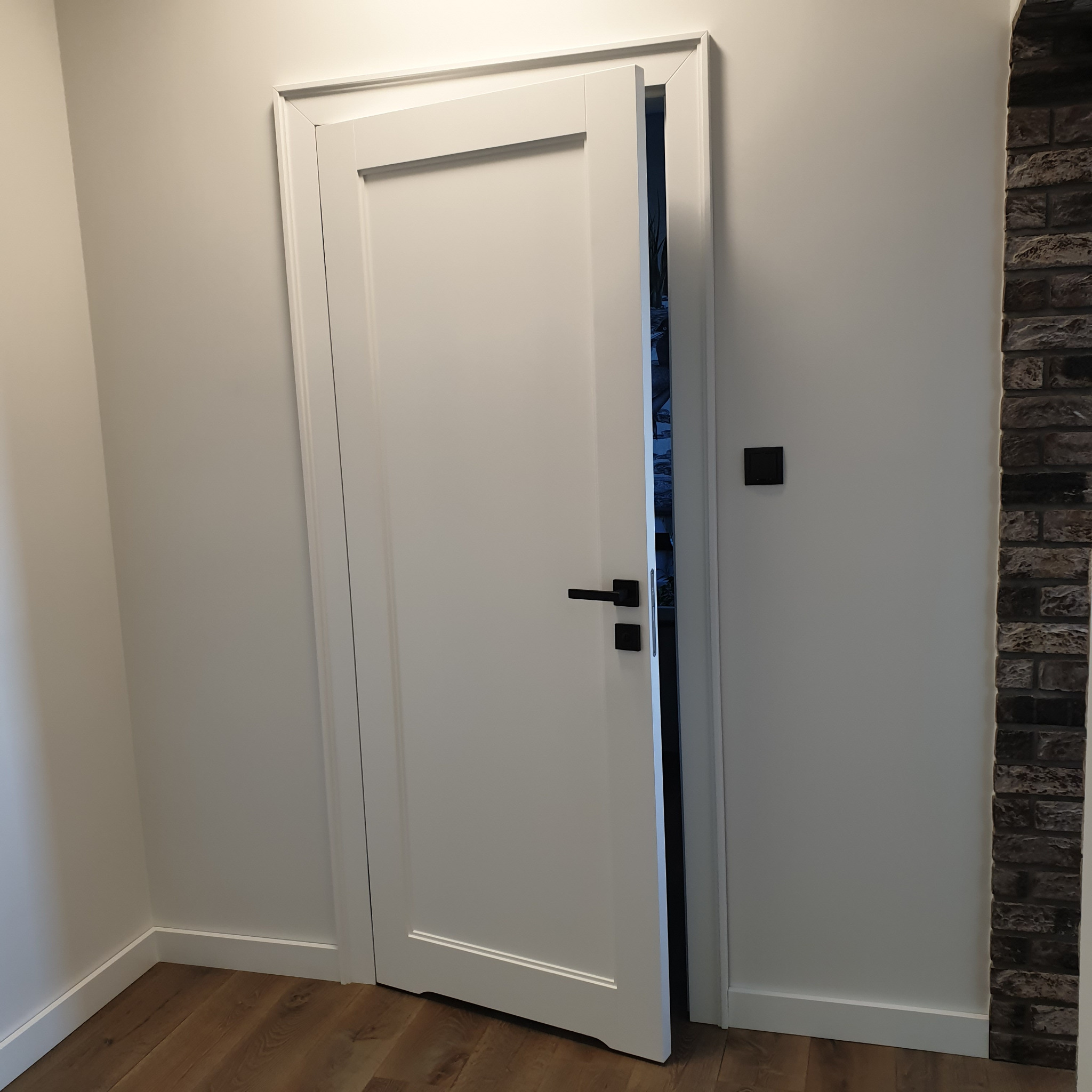 Białe, częściowo otwarte drzwi z czarną klamką, w minimalistycznym wnętrzu z drewnianą podłogą i fragmentem ściany wykończonej cegłą.