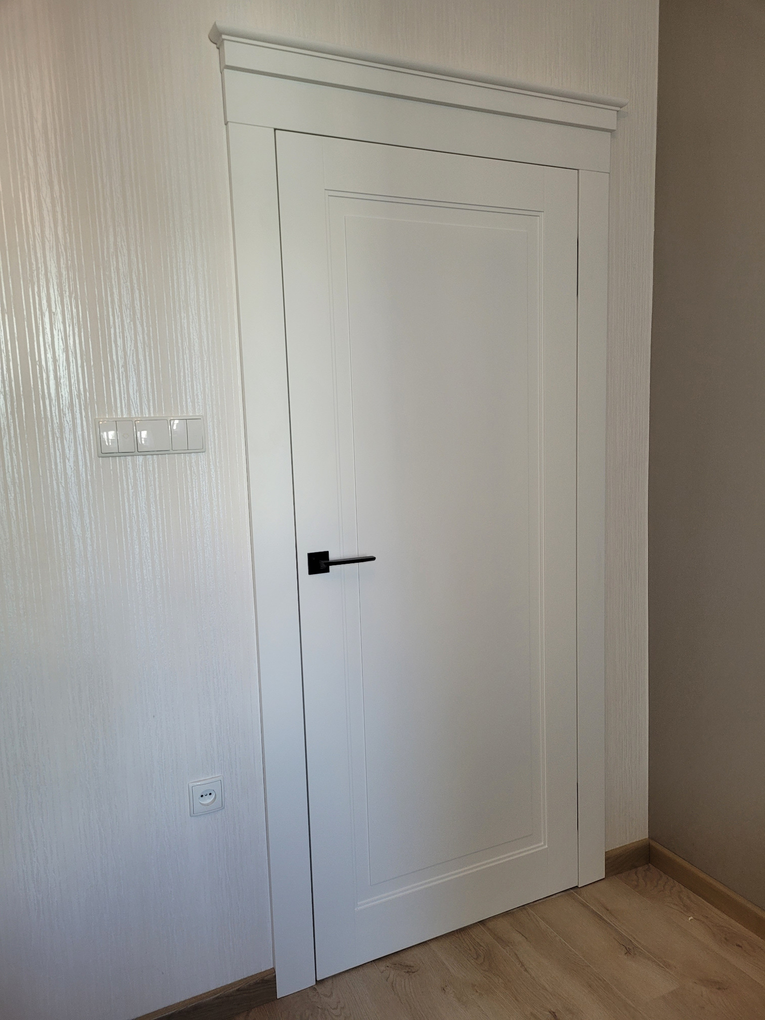 Białe, panelowe drzwi wewnętrzne z czarną klamką, osadzone w białej ościeżnicy. Na ścianie widoczny włącznik światła i gniazdko elektryczne. Podłoga drewniana.