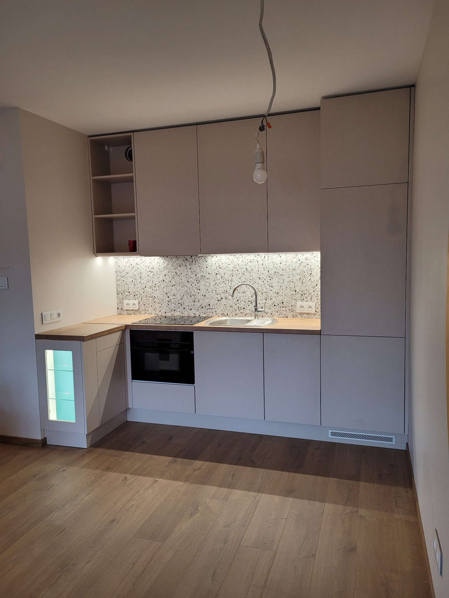 Jasna kuchnia na wymiar z drewnianym blatem i mozaiką nad zlewem. Widoczne oświetlenie podszafkowe i wisząca żarówka. Podłoga z paneli imitujących drewno.