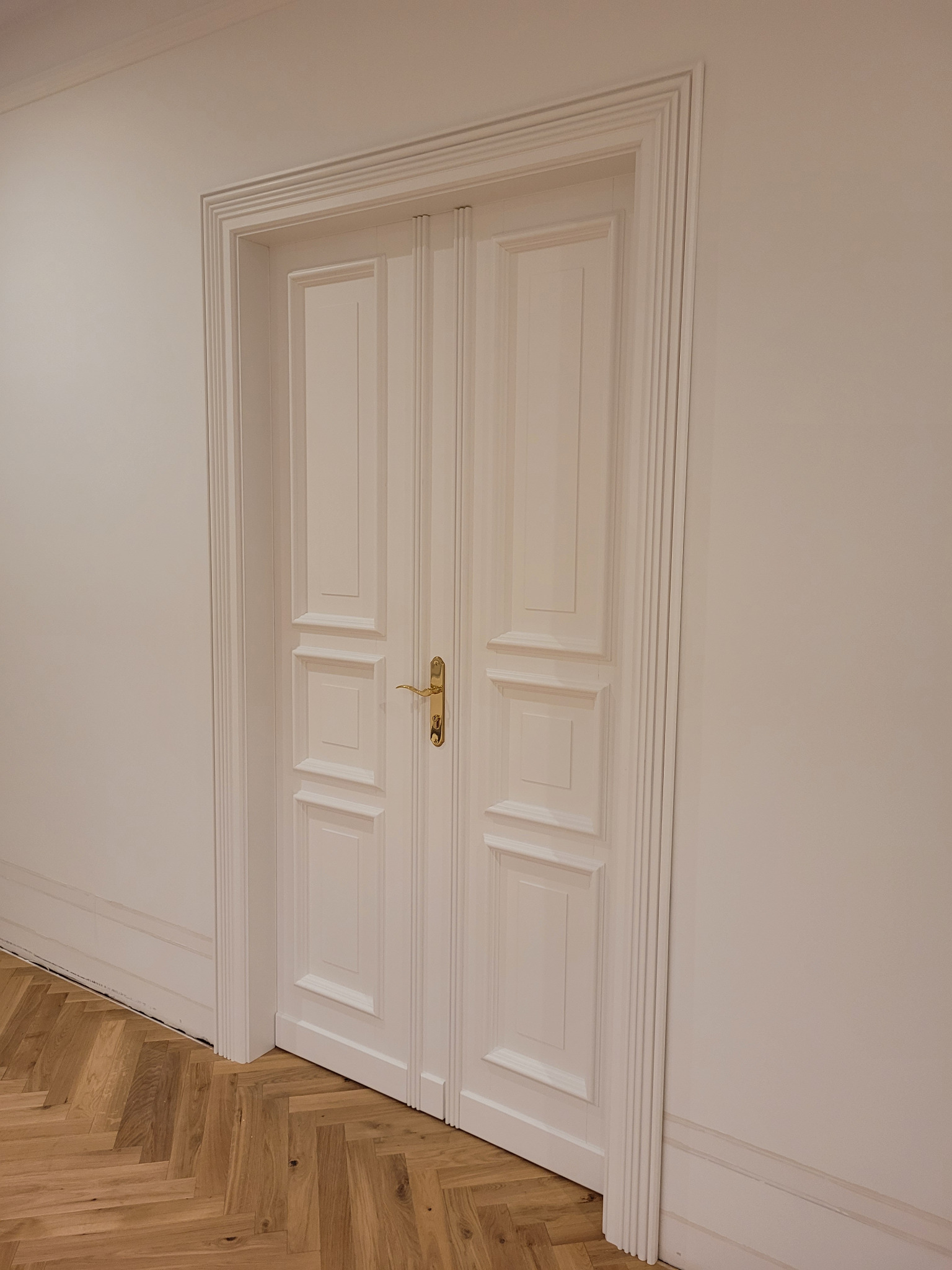 Białe, dwuskrzydłowe drzwi z ozdobnymi listwami i złotą klamką, widoczne na tle jasnej ściany i parkietu ułożonego w jodełkę.