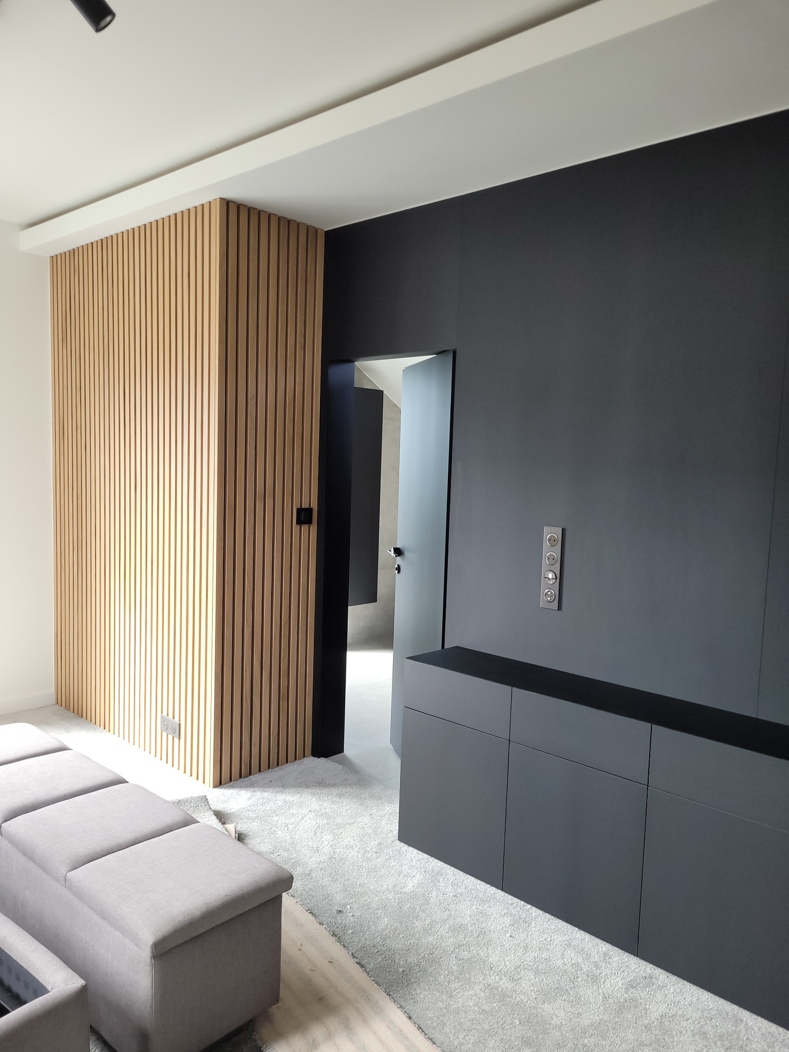 Nowoczesne wnętrze z drewnianą ścianką działową z lameli, czarną zabudową meblową i szarą sofą. Minimalistyczny design z dbałością o detale i oświetlenie.