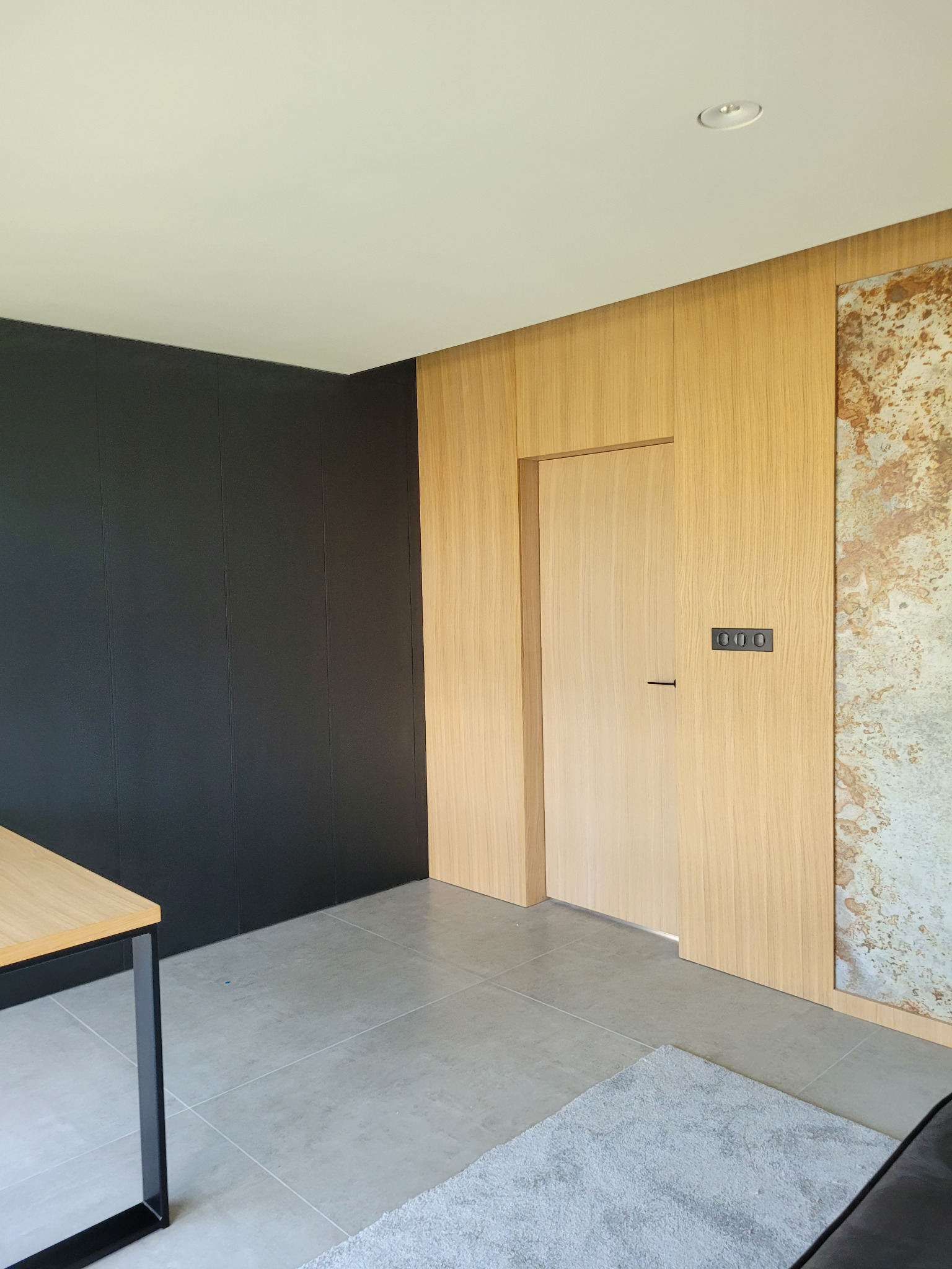 Nowoczesne wnętrze z ukrytymi drzwiami z jasnego drewna, wkomponowanymi w boazerię. Minimalistyczny design z czarnymi akcentami i szarą podłogą.