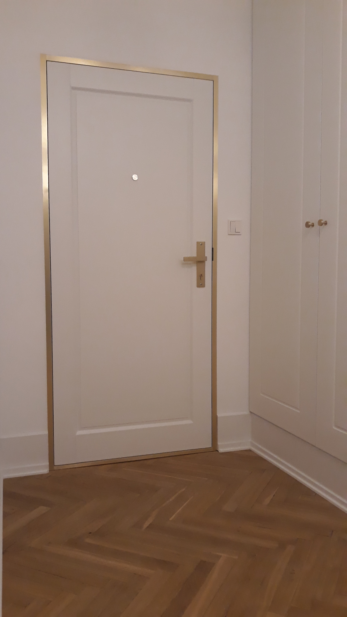 Białe drzwi z złotą ramką i wizjerem, zamontowane w białej ścianie, widoczne fragmenty parkietu i szafy wnękowej. Minimalistyczny design.