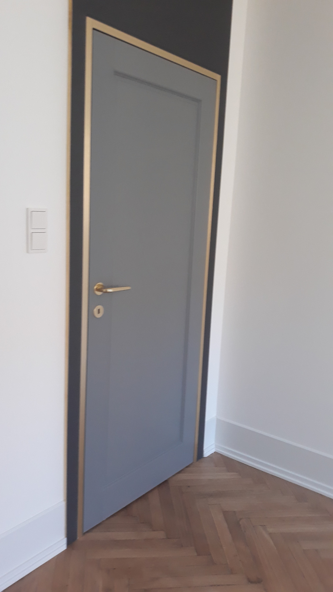 Szare, klasyczne skrzydło z złotą obwódką i klamką, zamontowane w białej ścianie. Na podłodze parkiet ułożony w jodełkę. Widoczny fragment ściany w ciemnym kolorze.