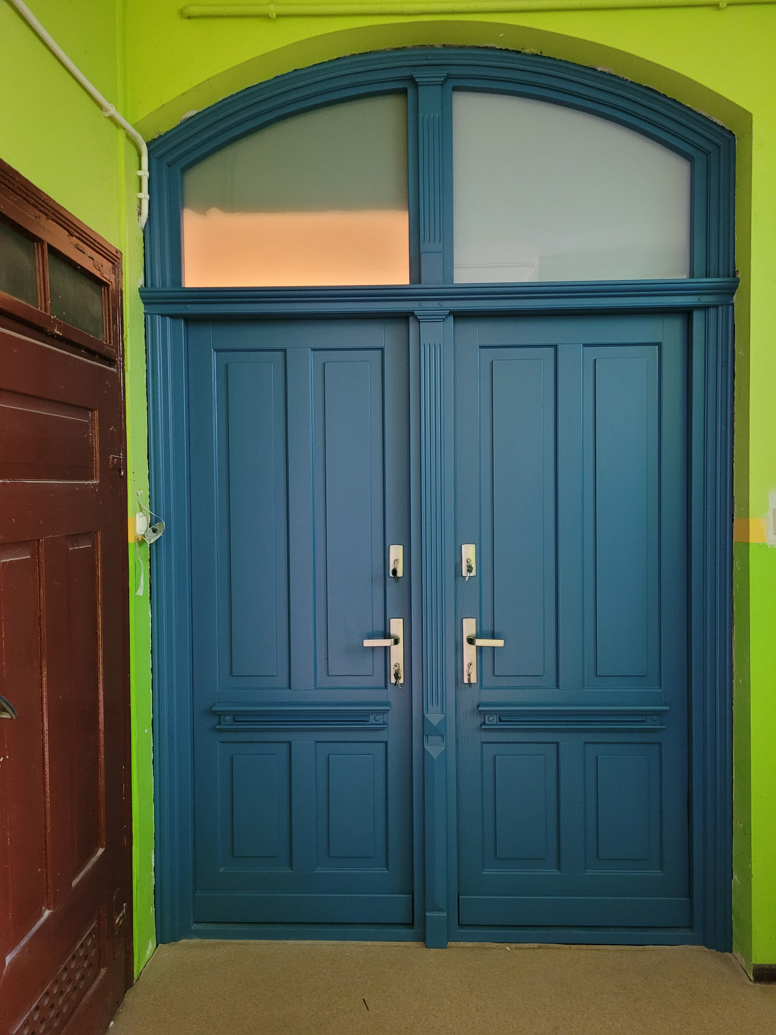 Dwuskrzydłowe, niebieskie drzwi z górnym naświetlem w łukowatej ramie, z widocznymi srebrnymi klamkami i kluczami, na tle limonkowo-zielonej ściany.
