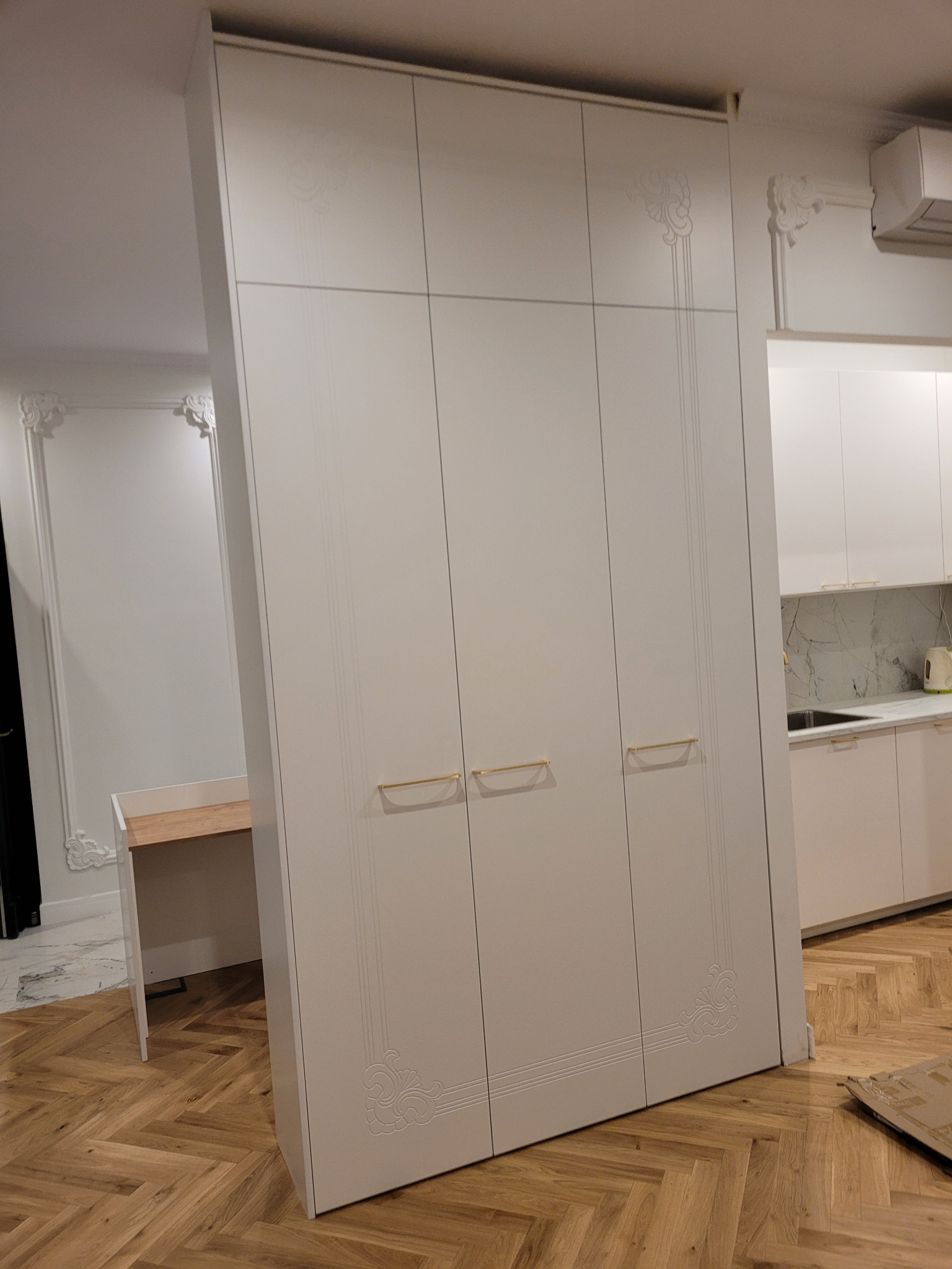 Biała szafa wnękowa z dekoracyjnymi frezami i złotymi uchwytami, wkomponowana w aranżację mieszkania w Buku. Widoczna zabudowa kuchenna w tle.