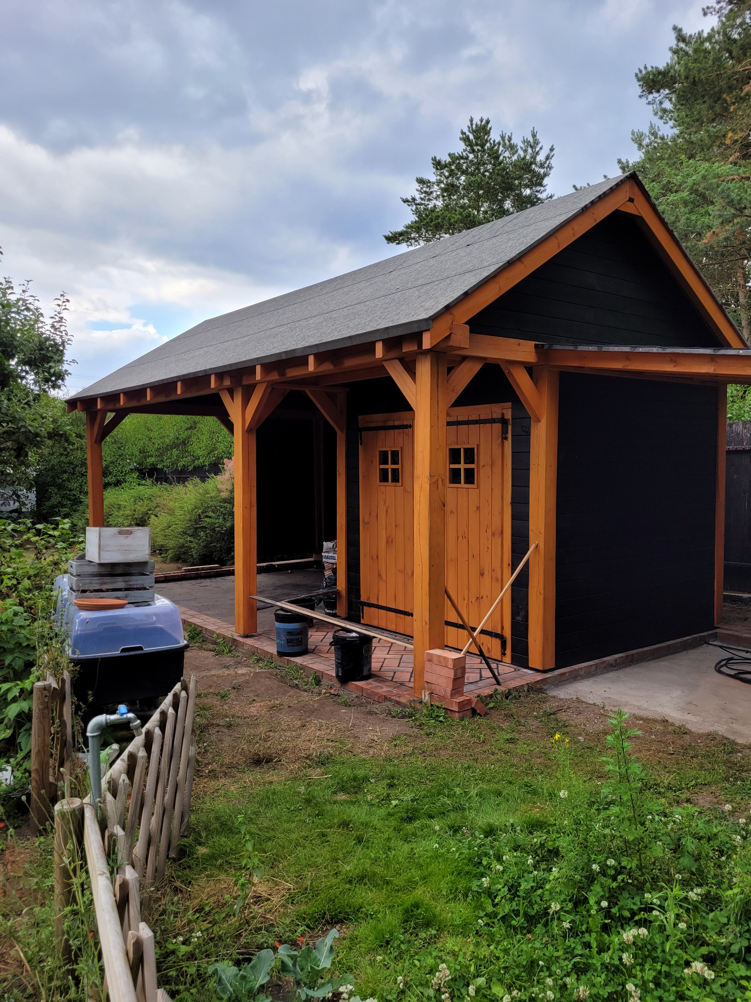 Drewniana altana z dwuskrzydłowymi drzwiami i zadaszeniem, pomalowana na czarno z elementami w kolorze naturalnego drewna. Widoczne narzędzia i materiały budowlane.