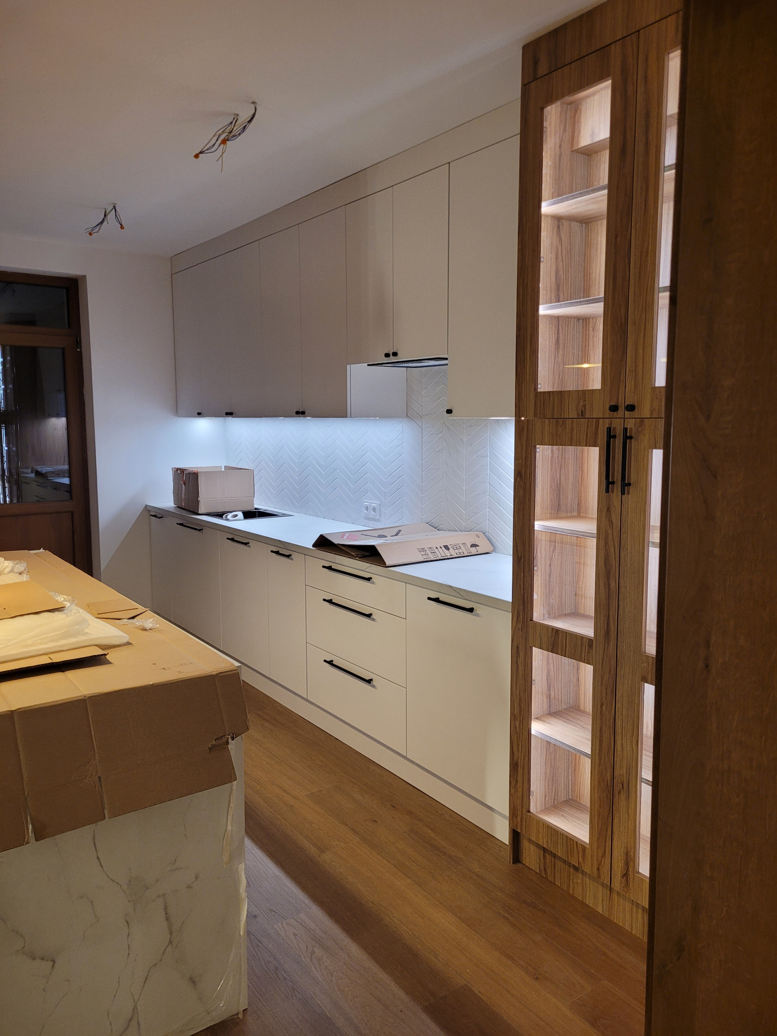 Nowoczesna, jasna kuchnia z białymi szafkami i drewnianą witryną z podświetleniem. Na blacie kartony, sugerujące świeży montaż. Uwagę przykuwa wzór jodełki na ścianie.