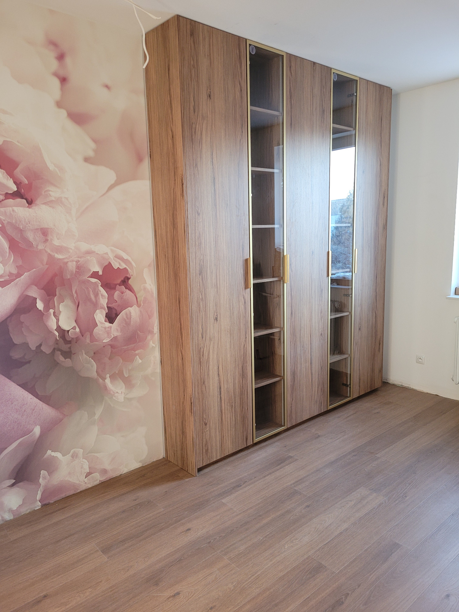 Wysoka, drewniana szafa wnękowa z przeszklonymi segmentami, złote obramowania, na tle ściany z motywem kwiatowym, parkiet na podłodze.