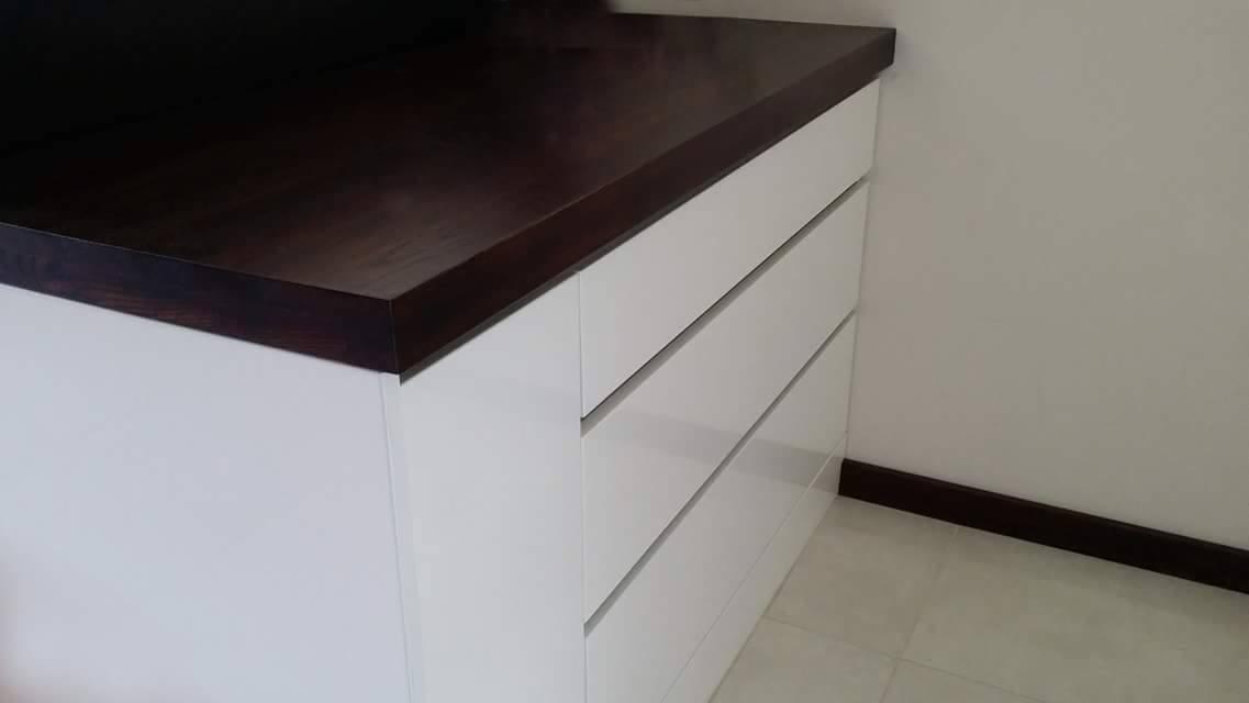 Biała komoda z trzema szufladami i blatem z ciemnego drewna, umieszczona w rogu pomieszczenia z jasnymi ścianami i podłogą.