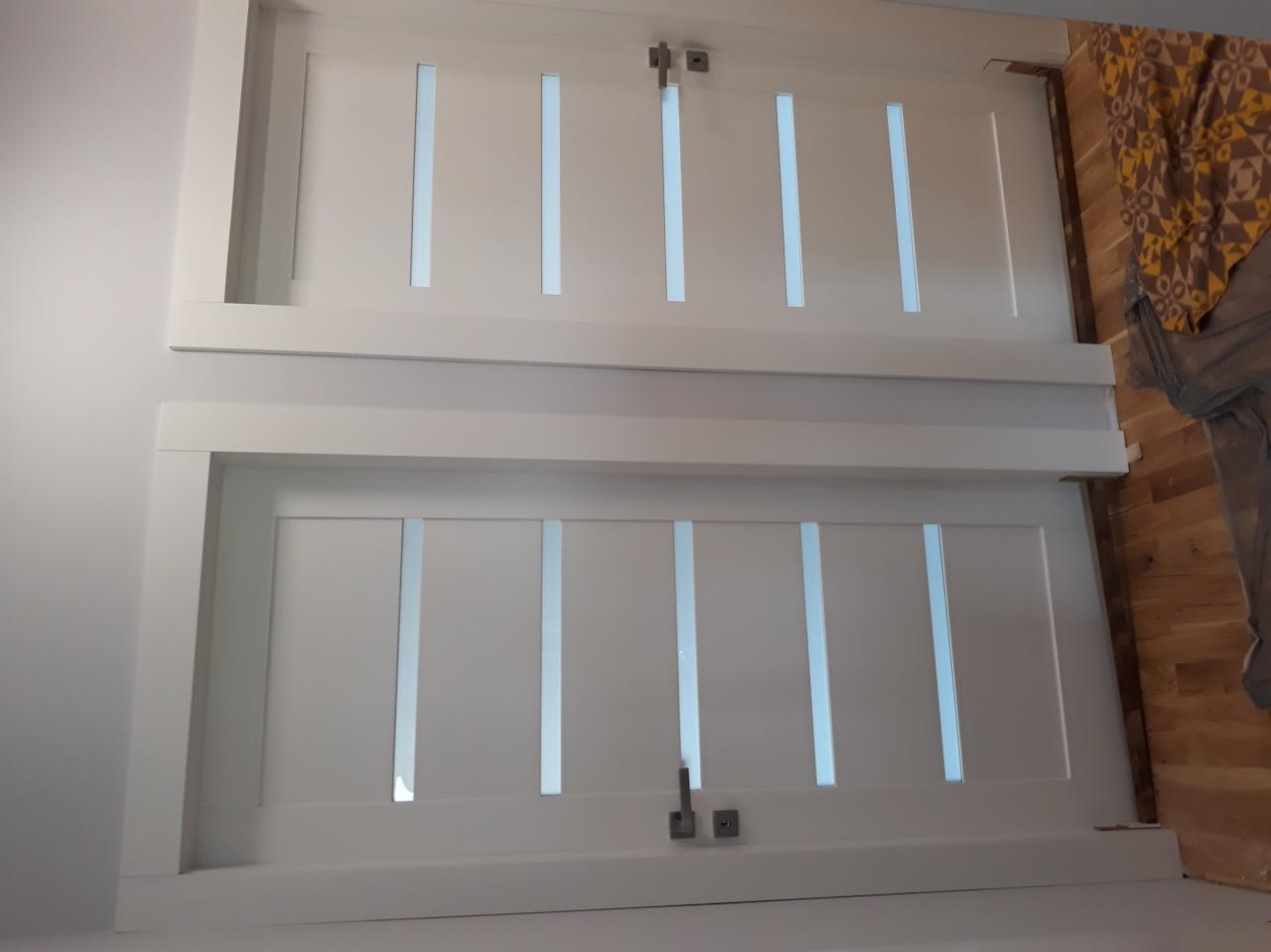 Dwa białe skrzydła drzwiowe z przeszkleniami w pionowe pasy, widoczne klamki i fragment podłogi w tle.