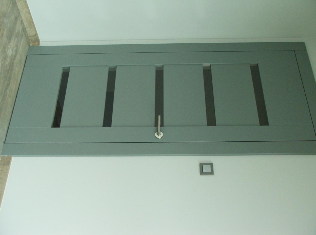 Szara szafka z przesuwnymi drzwiczkami z podłużnymi wycięciami, zamontowana wysoko na białej ścianie, widoczny metalowy uchwyt i włącznik światła.