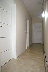 Korytarz z trzema białymi skrzydłami drzwiowymi o nowoczesnym designie, jasna podłoga z paneli, beżowe ściany, minimalistyczne oświetlenie sufitowe.