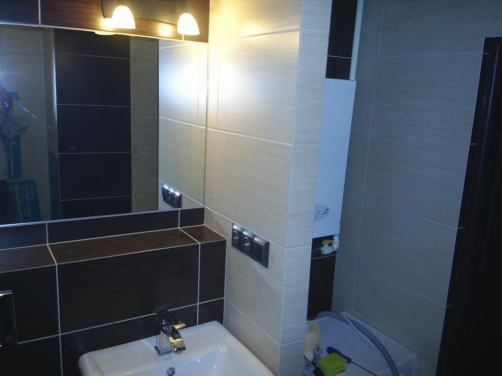 Wykończona łazienka z umywalką, lustrem, brązowymi i beżowymi płytkami, oraz piecem gazowym na ścianie.