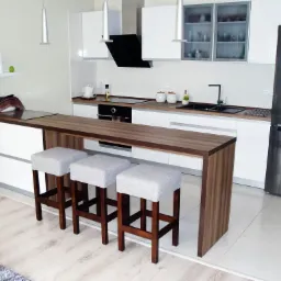 Nowoczesna, jasna kuchnia z białymi szafkami, drewnianym blatem i wyspą z trzema szarymi stołkami barowymi; widoczny srebrny, dwudrzwiowy lodówka i czarny okap.