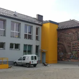 Budynek po remoncie z muralem przedstawiającym bibliotekę. Widoczny fragment dachu, okna, rampa dla niepełnosprawnych i samochód dostawczy.