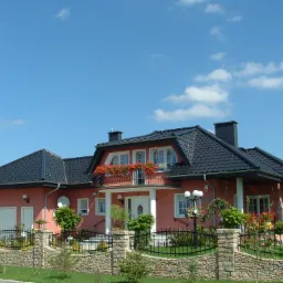 Dwukondygnacyjny dom z ciemną dachówką, balkonem z kwiatami i kamiennym ogrodzeniem, na tle błękitnego nieba.