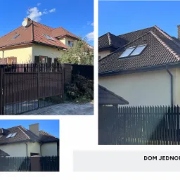 Żółty dom jednorodzinny z brązowym dachem, ceglanym kominem i brązowymi okiennicami, widoczny zza ciemnego, metalowego ogrodzenia.