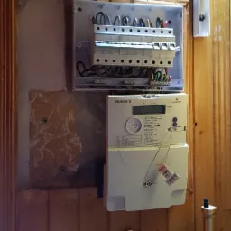 Zamontowana skrzynka z bezpiecznikami i licznik energii elektrycznej NORAX 3 na drewnianej ścianie, widoczne przewody i fragment kalendarza z portretem dziecka.