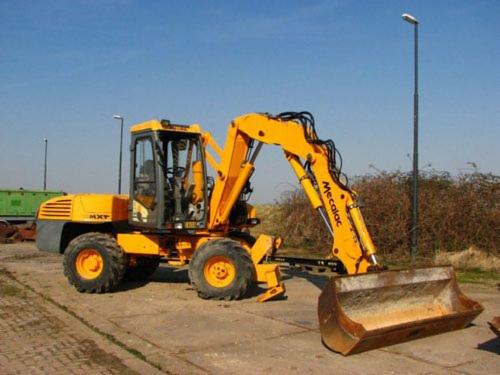 Żółta koparko-ładowarka Mecalac MXT na betonowym placu, z łyżką skierowaną w stronę widza, na tle nieba i zarośli.