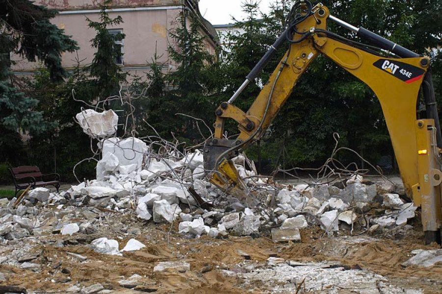 Żółta koparka CAT 434E kruszy gruz na placu budowy z ławką w tle, widoczne fragmenty zbrojenia.