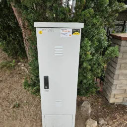 Biała szafka telekomunikacyjna z naklejkami informacyjnymi i oznaczeniami ostrzegawczymi, umieszczona na zewnątrz obok drzewa iglastego i ceglanego murku.