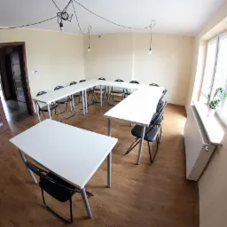 Sala szkoleniowa z białymi stołami ustawionymi w kształcie podkowy, czarnymi krzesłami, drewnianą podłogą i drzwiami z poziomymi paskami, oświetlona trzema wiszącymi żarówkami na kablach.