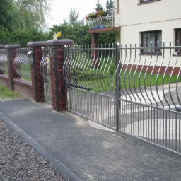 Nowoczesna brama wjazdowa ze stali nierdzewnej, z falistymi prętami, osadzona między ceglanymi słupkami. Widoczny fragment chodnika z kostki brukowej i fragment ogrodzenia z identycznymi przęsłami.