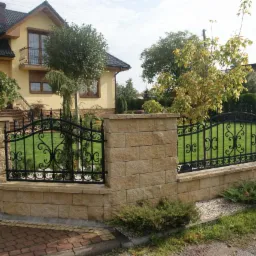 Eleganckie ogrodzenie z czarnymi, kutymi przęsłami i słupkami z piaskowca, otaczające zadbany trawnik przed domem z balkonem.