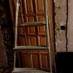 Tom-Bud.Uslugi-ogolnobudowlane - Stara, drewniana, brązowa futryna drzwiowa oparta o ścianę, obok stoi aluminiowa drabina. W tle surowa ściana z widocznymi śladami prac remontowych.