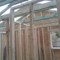 Tom-Bud.Uslugi-ogolnobudowlane - Szkieletowa konstrukcja domu w trakcie budowy, widoczne drewniane ramy ścian i stropu, płyty OSB na ścianach, okno z widokiem na zewnątrz. Konstrukcja w surowym stanie.