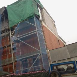 Tom-Bud.Uslugi-ogolnobudowlane - Budowa domu z czerwonej cegły, widoczne rusztowania z siatką ochronną i plandeką. Na dole fragment samochodu dostawczego.