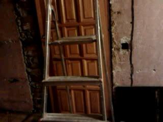 Stara, drewniana, brązowa futryna drzwiowa oparta o ścianę, obok stoi aluminiowa drabina. W tle surowa ściana z widocznymi śladami prac remontowych.