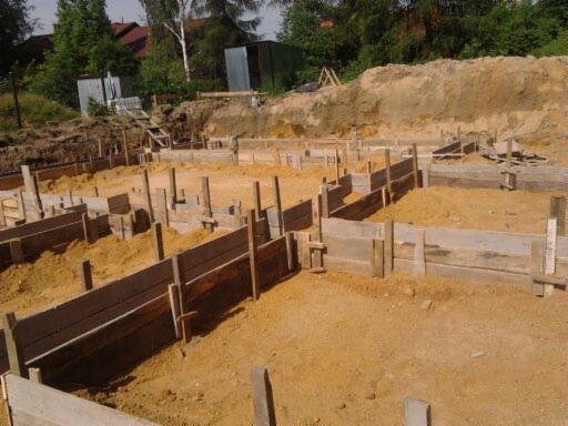 Deskowanie fundamentów domu jednorodzinnego z widocznym wykopem i elementami konstrukcyjnymi. Prace ziemne w toku, widok z góry na plac budowy.