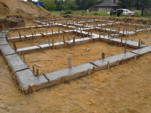 Wylane fundamenty domu jednorodzinnego, z widocznym szalunkiem i świeżym betonem. Na drugim planie hałda ziemi i dom w budowie.