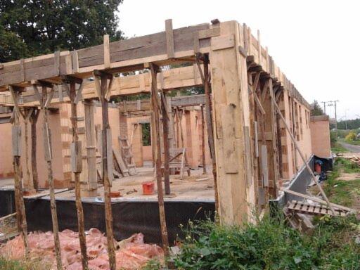 Wznoszenie ścian domu z cegły z drewnianym szalunkiem. Widoczne elementy konstrukcyjne i teren budowy. Ujęcie z poziomu gruntu, z zielenią w kadrze.