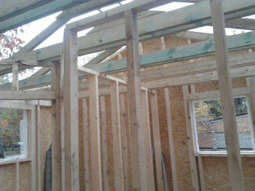 Szkieletowa konstrukcja domu w trakcie budowy, widoczne drewniane ramy ścian i stropu, płyty OSB na ścianach, okno z widokiem na zewnątrz. Konstrukcja w surowym stanie.