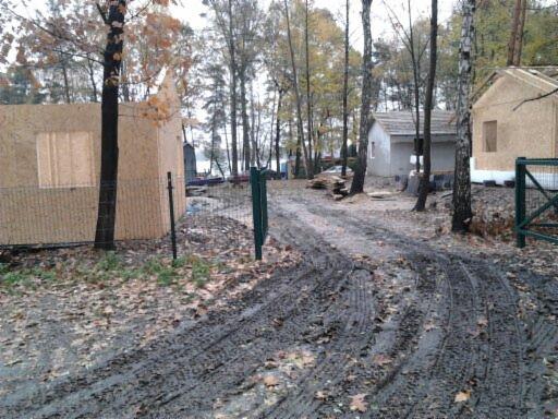 Wczesna faza budowy domu szkieletowego z widoczną konstrukcją ścian i dachu, otoczonego drzewami i błotnistą drogą dojazdową w jesiennej scenerii.