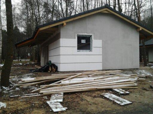 Niewielki, otynkowany dom w trakcie budowy, z częściowo ocieploną elewacją i stertą desek na ziemi. Widoczne drzewa w tle, pochmurna pogoda.