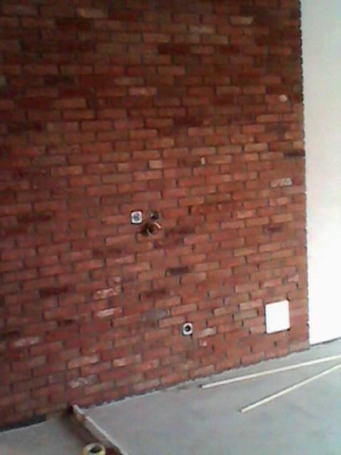 Ceglana ściana z widocznymi otworami na instalacje, surowy beton na podłodze i fragmenty rurek. Wnętrze w trakcie prac wykończeniowych.