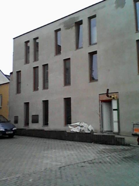 Surowy budynek z szarymi tynkami i rzędami okien, widoczne ślady napraw na elewacji. Przed budynkiem zaparkowany samochód i materiały budowlane.