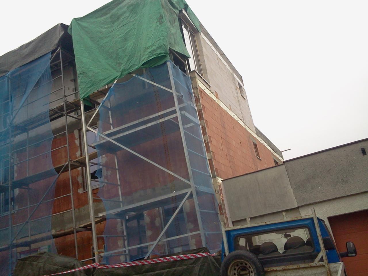 Budowa domu z czerwonej cegły, widoczne rusztowania z siatką ochronną i plandeką. Na dole fragment samochodu dostawczego.