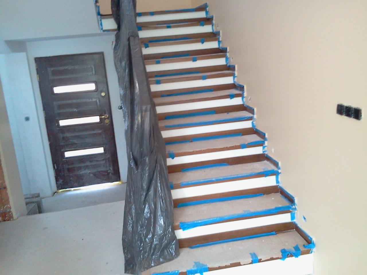Wnętrze w trakcie prac wykończeniowych: schody drewniane zabezpieczone taśmą malarską i folią ochronną, drzwi wewnętrzne z przeszkleniami w tle.
