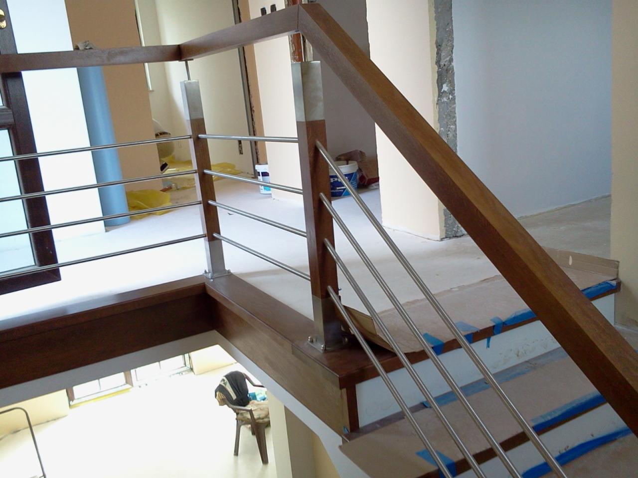Nowoczesne schody z drewnianą balustradą i metalowymi prętami w jasnym wnętrzu. Widoczne zabezpieczone schody i fragmenty prac wykończeniowych.