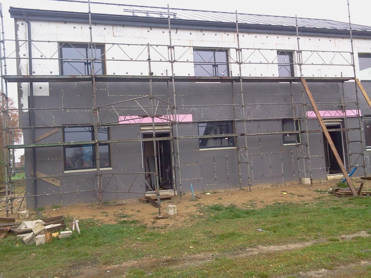 Budynek w trakcie ocieplania, widoczne rusztowania, szara izolacja termiczna na ścianach, biała izolacja na górze. Częściowo ocieplona elewacja z oknami.