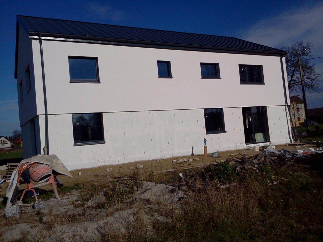 Dwukondygnacyjny dom w trakcie budowy, z oknami i ciemnym dachem. Część elewacji ocieplona, widoczna betoniarka i materiały budowlane na ziemi.